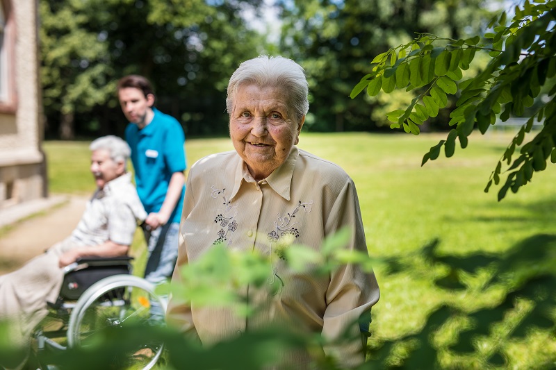 Eine ältere Frau lächelt in die Kamera, während im Hintergrund in einer sonnigen Parklandschaft eine Pflegekraft eine andere ältere Person im Rollstuhl schiebt.