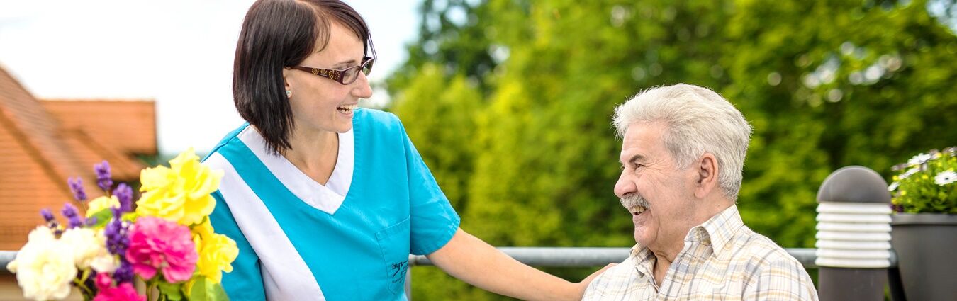 Eine Krankenschwester in blauer Uniform unterhält sich lächelnd mit einem älteren Mann, der im Freien sitzt, und bespricht Angebote für ältere Menschen.
