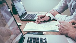 Zwei Personen sitzen vor Laptops und Schreibpapier, zu sehen sind nur die Hände und Arme. Beide halten Stifte in der Hand.