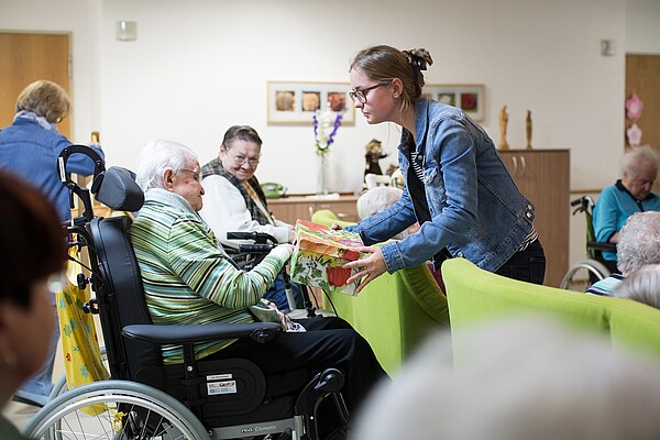 Jüngere Frau überreicht einer Seniorin, die im Rollstuhl sitzt, ein Geschenk. Im Hintergrund sind weitere Senioren und Seniorinnen zu sehen.