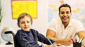 Junge im Rollstuhl sitzt mit einem Betreuer an einem Tisch.