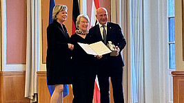 Drei Personen stehen auf einem Podest. In der Mitte und links stehen eine Frau, rechts ein Mann. Die Frau in der Mitte hält eine zweiseitiges Dokument vor sich. Im Hintergrund sind drei Flaggen. 