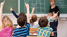 Schulkinder in einem Klassenzimmer. Alle Schüler*innen melden sich, um eine Frage zu beantworten. Im Hintergrund eine Lehrerin, die vor einer Tafel steht.