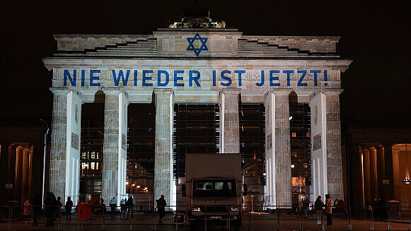 Brandenburger Tor im Dunkeln. Oben ist ein blauer Davidstern zusammen mit der Aufschrift "Nie wieder ist jetzt!" zu sehen. Vor dem Tor steht ein LkW, mehrere Passanten sind ebenfalls abgebildet.