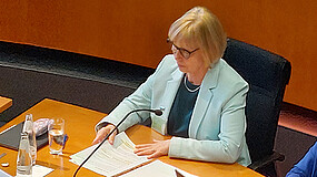 IB-Vorständin Karola Becker sitzt auf einem Stuhl im Deutschen Bundestag. Vor ihr liegt auf einem Tisch ein Stapel mit Papier.