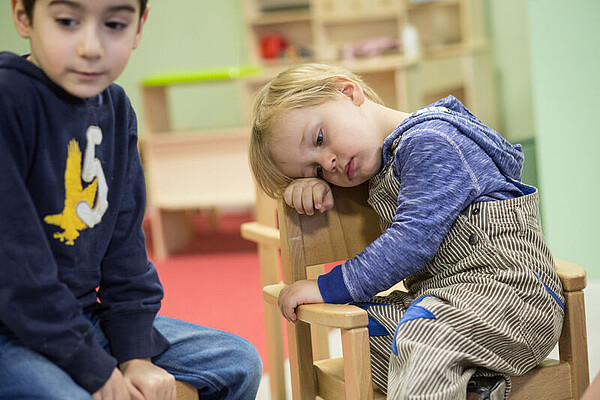 Zwei Kinder sitzen auf Stühlen in einer Kita. Ein Kind hat den Kopf auf die Lehne seines Stuhls gelegt.