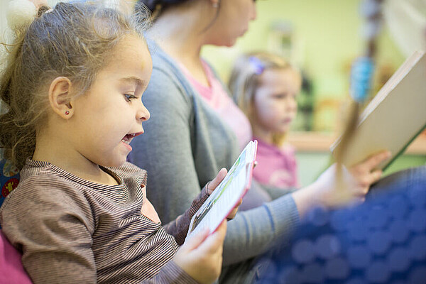 Kinder und Betreuerin sitzen in einer Kita nebeneinander und lesen aus Büchern vor.