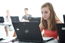 Junge Frau sitzt an einem Tisch vor einem Laptop. Im Hintergrund sind zwei weitere Personen zu sehen.
