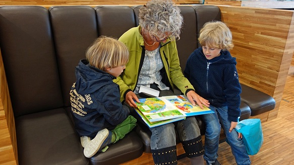 Ältere Person sitzt mit zwei Kindern auf einem Sofa und liest aus einem Buch vor.