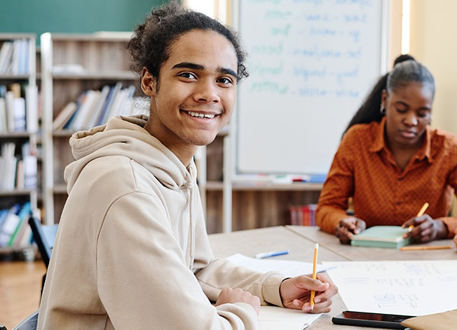 Zwei Personen sitzen an einem Tisch mit Schreibutensilien und Notizbüchern, im Hintergrund Bücherregale und ein Flipchart.