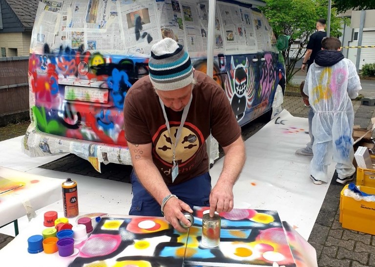 Projektschmiede_Spray-Aktion des Projekts "BusSTOP" Person mit Mütze sprüht bunte Graffiti auf eine große weiße Fläche vor einem mit Zeitungen abgedeckten Fahrzeug, weitere Personen im Hintergrund mit Schutzanzügen