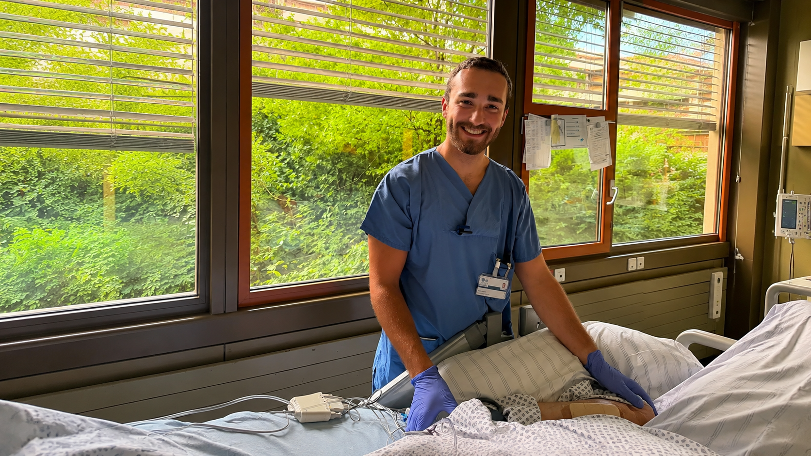 Malte steht lächelnd in blauer Pflegekleidung an einem Patientenbett auf einer Intensivstation.