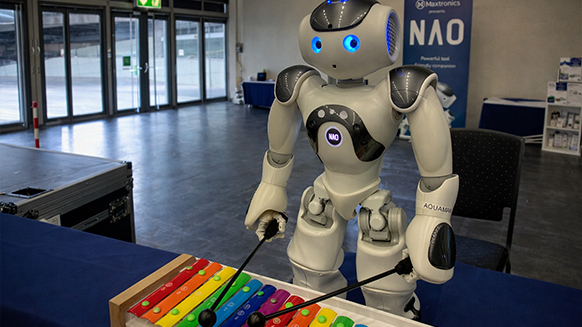Neu bei der Didacta 2026: ein stark ausgebauter Bereich zu den Themen Robotik und Künstliche Intelligenz. Foto: IB | KI-bearbeitet Ein weißer Roboter spielt auf einem bunten Xylophon. Im Hintergrund ist eine Halle.