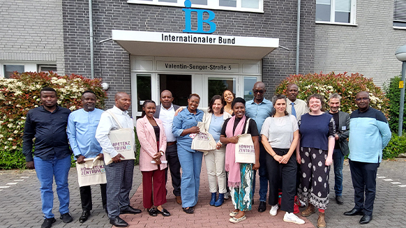 15 Personen mit schwarzer und weißer Hautfarbe stehen vor dem Haupteingang der Zentralen Geschäftsführung des IB. Der Eingang besteht aus einer weißgerahmten Glastür, beschriftet mit "Internationaler Bund" und "Valentin-Senger-Straße 5". Über der Tür befindet sich ein blaues IB-Logo.