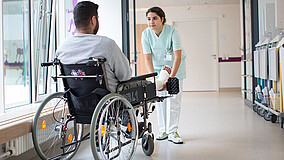 Mann mit Gips sitzt im Rollstuhl, vor ihm steht eine Krankenschwester