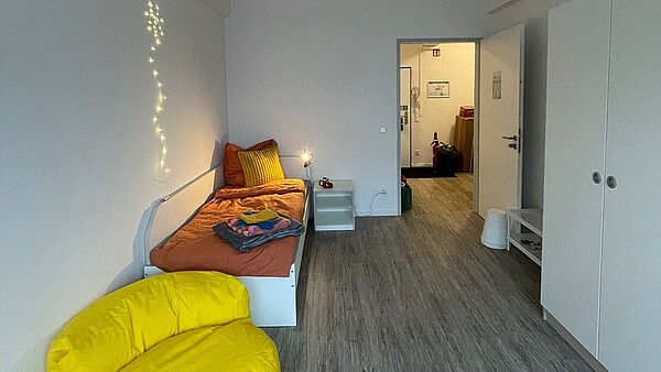 Ein Zimmer der Einrichtung mit gelber Couch, Bett und einer Lichterkette an der Wand.