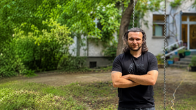 Arian Begovic steht vor dem Gebäude der Hitzehilfe des Internationalen Bundes (IB) in Berlin. Vor der Einrichtung sind Grünflächen und Bäume zu sehen.