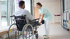 Pflegekraft in hellgrünem Kittel beugt sich zu einem Mann im Rollstuhl in einem Krankenhausflur hinunter.