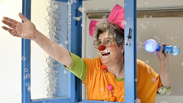 Nicole Hornberger als Clownin mit Clownsschminke lächelt aus einem Fenster, während Seifenblasen durch die Luft schweben.