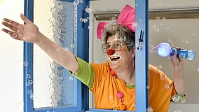 Nicole Hornberger als Clownin mit Clownsschminke lächelt aus einem Fenster, während Seifenblasen durch die Luft schweben.