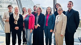 Mehrere Personen stehen zusammen, hinter ihnen ist die Glaskuppel des Deutschen Bundestags in Berlin zu erkennen.