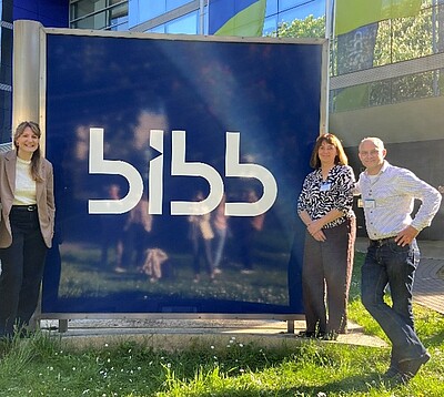 Drei Personen stehen neben einem großen blauen Schild mit weißer Aufschrift 'bibb' vor einem modernen Gebäude.