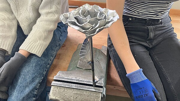 Metallrose in einem Schraubstock zwischen zwei sitzenden Personen mit Arbeitshandschuhen