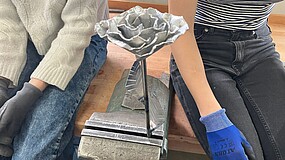 Metallrose in einem Schraubstock zwischen zwei sitzenden Personen mit Arbeitshandschuhen