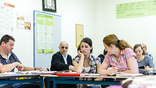Teilnehmerinnen und Teilnehmer eines Sprachkurses beim gemeinsamen Lernen in der Unterrichtssituation. Im Hintergrund sind Lernmaterialien an der Wand zu sehen.