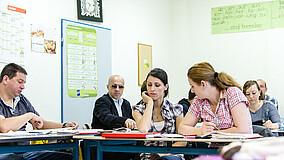 Teilnehmerinnen und Teilnehmer eines Sprachkurses beim gemeinsamen Lernen in der Unterrichtssituation. Im Hintergrund sind Lernmaterialien an der Wand zu sehen.