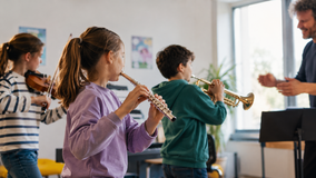 Drei Kinder spielen Geige, Querflöte und Trompete in einem hellen Raum, ein Erwachsener dirigiert sie.