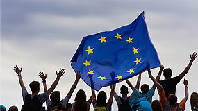 Eine Gruppe junger Menschen von hinten. Die Personen halten zusammen eine große Europa-Flagge hoch.