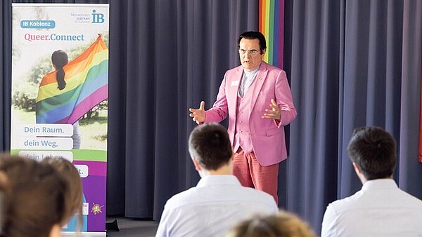 Thiemo Fojkar steht auf einer Bühne und hält eine Ansprache zur Eröffnung der Beratungsstelle "Queer.Connect". Er trägt ein rosafarbenes Jackett. Auf der Bühne steht ein Aufsteller mit Informationen zur Beratungsstelle. Ein Teil des Publikums ist von hinten abgebildet.