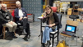 Sterenn Coudray sitzt auf einem Stuhl neben einem geöffneten Laptop; zwei Männer sind ebenfalls im Raum zu sehen.