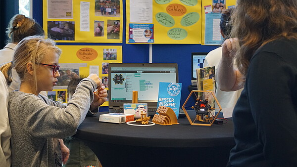 Kind sitzt an einem runden Tisch mit Laptop, kleinen Robotern und Informationsmaterial, im Hintergrund bunte Pinnwand mit Texten und Fotos.