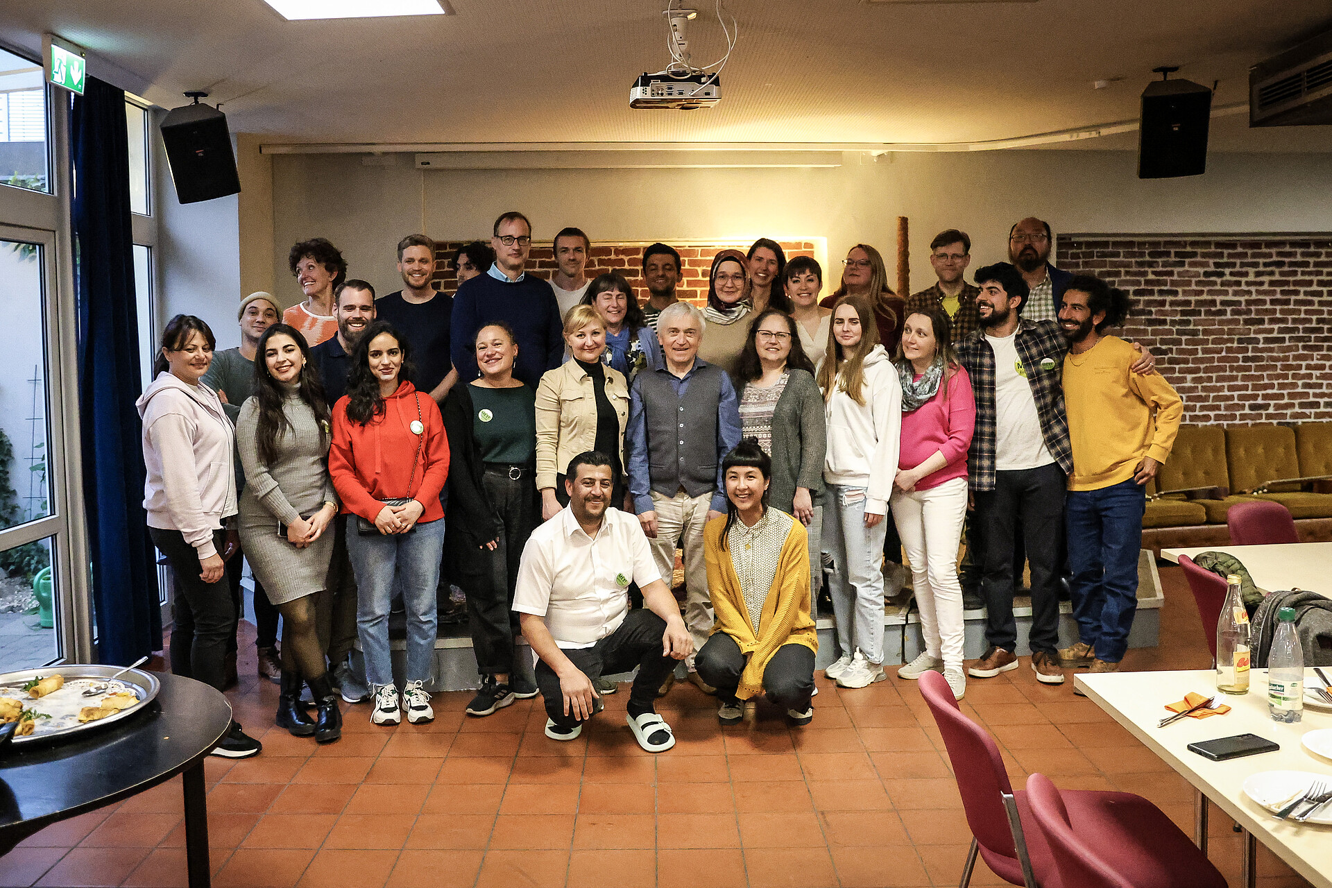 Projektschmiede_Das BAMF zu Besuch im IB Bamberg Gruppe von Menschen in einem Raum mit Backsteinwand und Sitzgelegenheiten, einige sitzen, andere stehen, im Vordergrund Tische mit Getränken und Tellern
