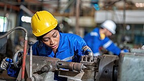 Zwei Personen in Schutzhelmen und Arbeitskleidung bedienen eine Maschine in einer Werkstatt oder Fabrikhalle