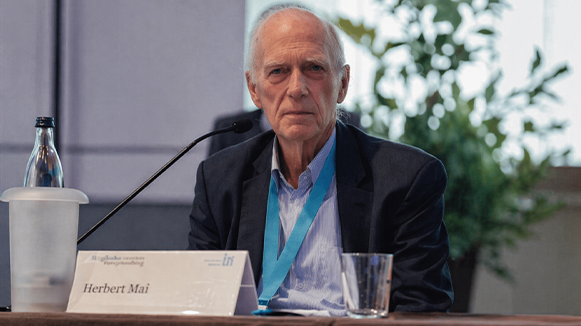 Herbert Mai sitzt an einem Rednerpult, vor ihm befindet sich ein Mikrofon. Auf dem Pult steht ein Schild, auf dem sein Name zu lesen ist. Im Hintergrund ist ein Fenster und davor eíne Zimmerpflanze.