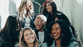 Sechs Personen sitzen auf einer Treppe, tragen lässige Kleidung, darunter Jeansjacken und Pullover, und blicken in verschiedene Richtungen.