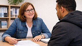 Ein Mann mit dunklem Teint ist von hinten zu sehen: Er sitzt an einem Schreibtisch einer Frau gegenüber. Die Frau lächelt. Vor ihr liegen Unterlagen, in der Hand hält sie einen Stift. Im Hintergrund ist ein Regal mit Ordnern und einer Zimmerpflanze.