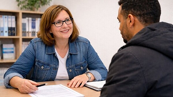 Ein Mann mit dunklem Teint ist von hinten zu sehen: Er sitzt an einem Schreibtisch einer Frau gegenüber. Die Frau lächelt. Vor ihr liegen Unterlagen, in der Hand hält sie einen Stift. Im Hintergrund ist ein Regal mit Ordnern und einer Zimmerpflanze.