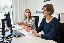 Zwei Frauen sitzen in einem Büro an einem Schreibtisch und schauen auf einen Monitor. Sie unterhalten sich.