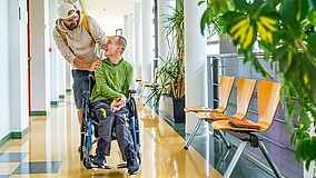 Ein Mann schiebt einen Jungen im Rollstuhl; beide lächeln sich an.