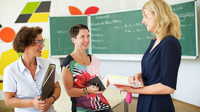 Drei Frauen befinden sich in einem Klassenzimmer. Zwei lehnen an einem Tisch, die dritte Frau steht daneben. In den Händen halten sie Ordner und Unterlagen. Im Hintergrund ist eine Tafel und eine bunt bemalte Wand zu sehen.