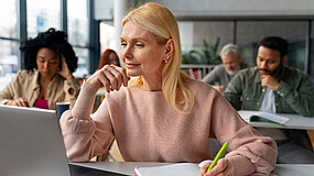 Eine Frau sitzt im Vordergrund an einem Laptop und macht sich Notizen. Im Hintergrund arbeiten weitere erwachsene Teilnehmende an Computern in einem Schulungsraum.