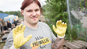 Ein junger Mann steht im Freien auf einem erdigen Boden: Er trägt ein hellgraues T-Shirt mit der Aufschrift "Ich bin freiwillig hier". Er hält die Hände etwas höher und trägt gelbe Arbeitshandschuhe. Im Hintergrund sind Büsche und weitere Personen zu sehen.