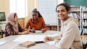 Drei junge Erwachsene sitzen in einem Unterrichtsraum eines Sprachkurses. Ein junger Mann und zwei junge Frauen arbeiten gemeinsam im Kurs, eine Person blickt lächelnd in die Kamera.
