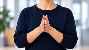Eine Person steht in einem Raum. Abgebildet ist nur der Oberkörper. Die Person presst ihre beiden Handflächen gegeneinander. Sie trägt einen dunkelblauen Pullover. Der Hintergrund ist hell und verschwommen.