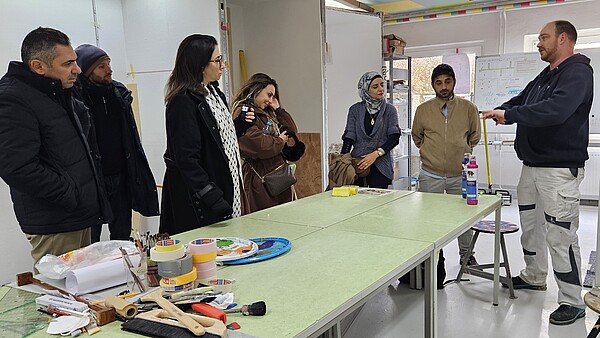 Ein Mann in Arbeitskleidung zeigt einer EVBB-Delegation Malerutensilien auf einem Arbeitstisch in der Werkstatt des Bildungscampus Neuenhagen des Internationalen Bundes