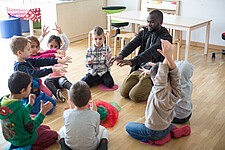 Mehrere Kinder sitzen in einer Kita zusammen mit einem Betreuer auf dem Boden uns spielen.